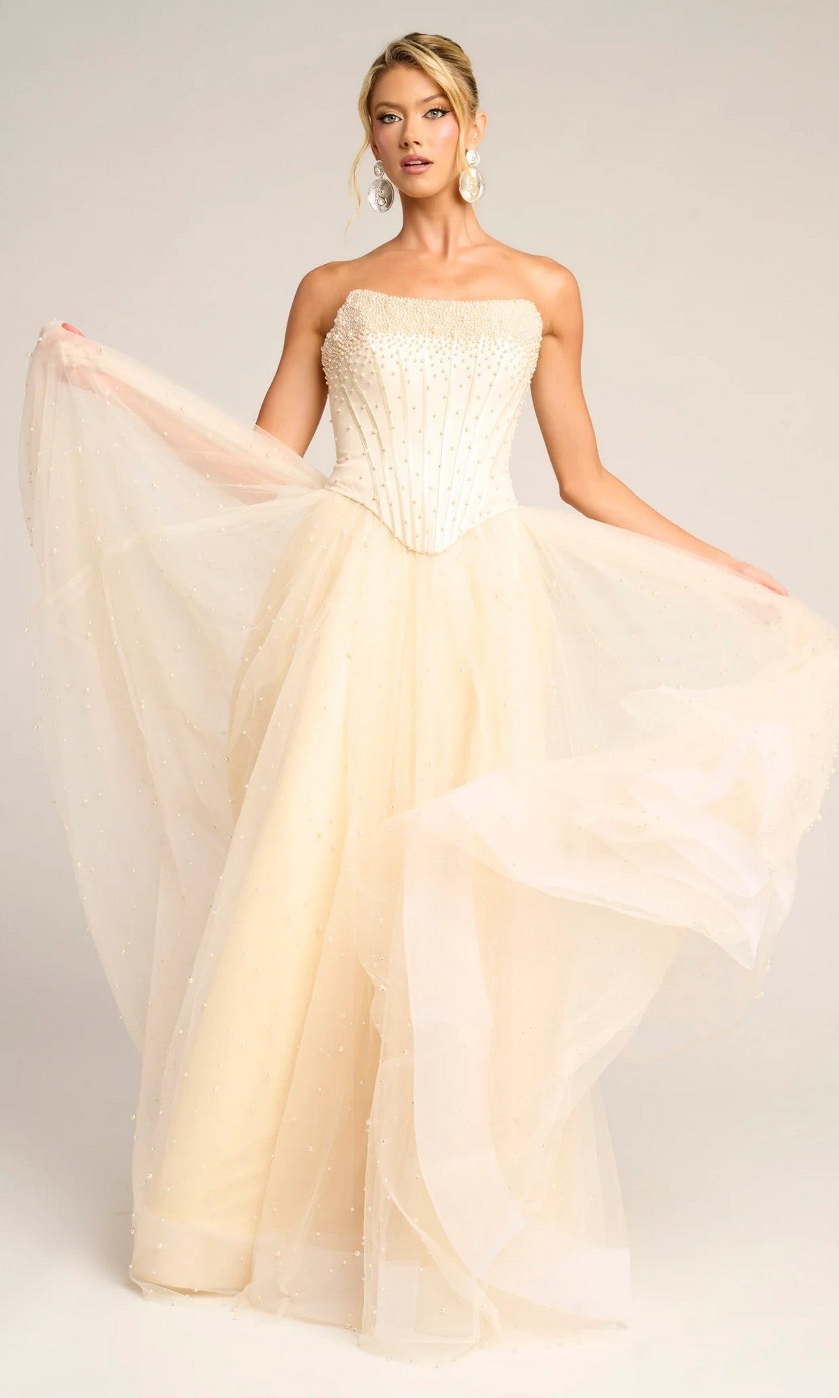 A woman stands holding the sides of her Portia and Scarlett PS26034 long formal dress in cream, featuring a strapless beaded corset bodice and a full glitter tulle skirt.